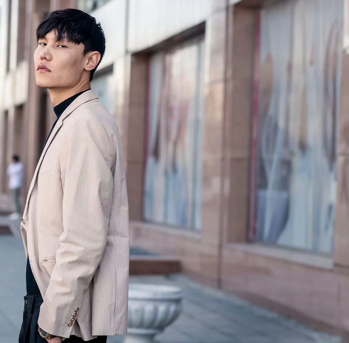 A man with dark hair, dressed in a beige blazer and black turtleneck, standing outdoors near a building with large windows - Acne Treatment for men at HIM Plastic Surgery