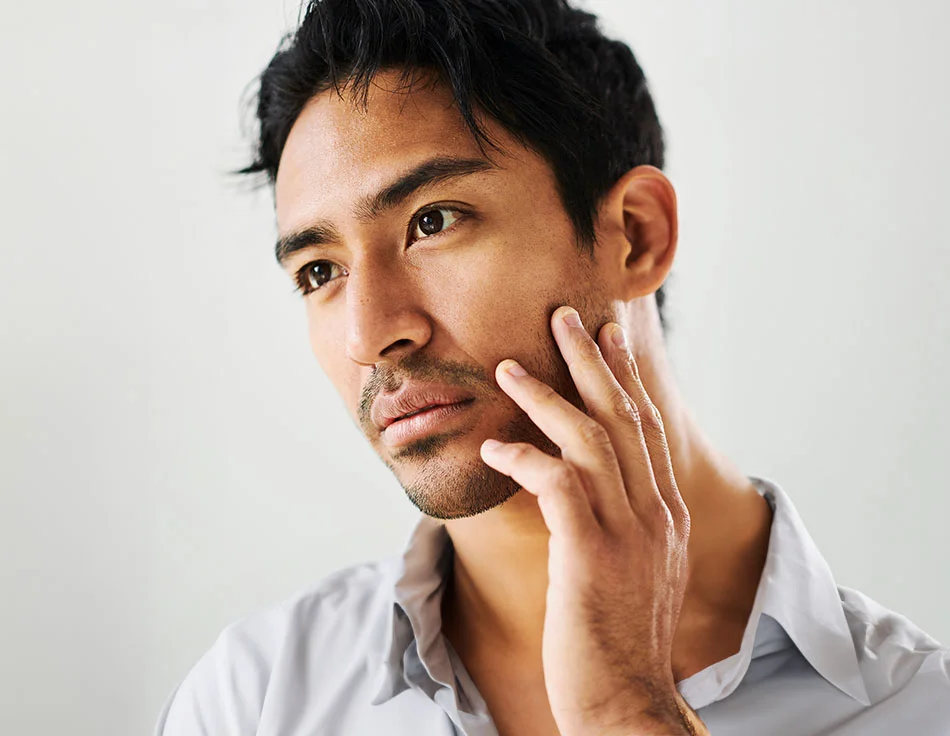 A close-up of a man touching his face, looking pensive, with a light background - Acne Treatment for men at HIM Plastic Surgery