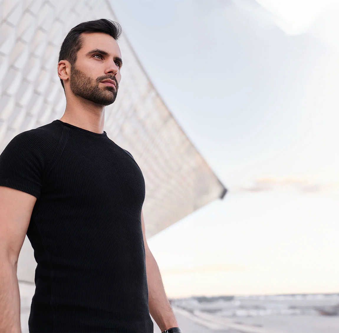 A man with dark hair and a beard, wearing a black t-shirt, standing outdoors with a modern building in the background - Botox for men at HIM Plastic Surgery