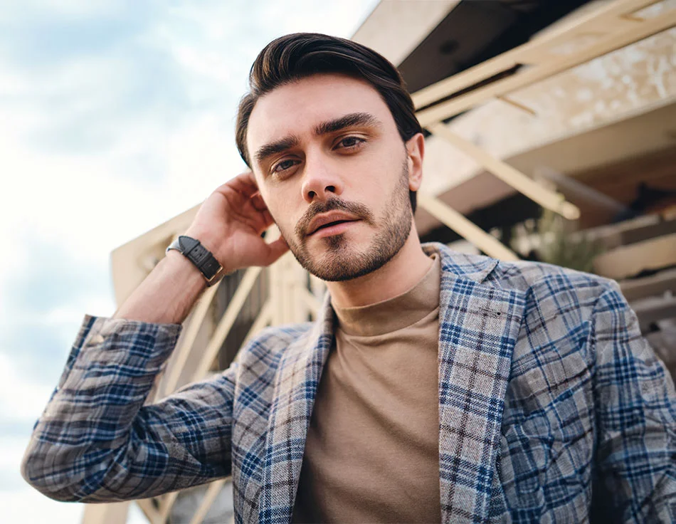 A man with neatly styled hair and a beard, wearing a beige turtleneck and a plaid blazer, standing outdoors with a building behind him - Botox for men at HIM Plastic Surgery