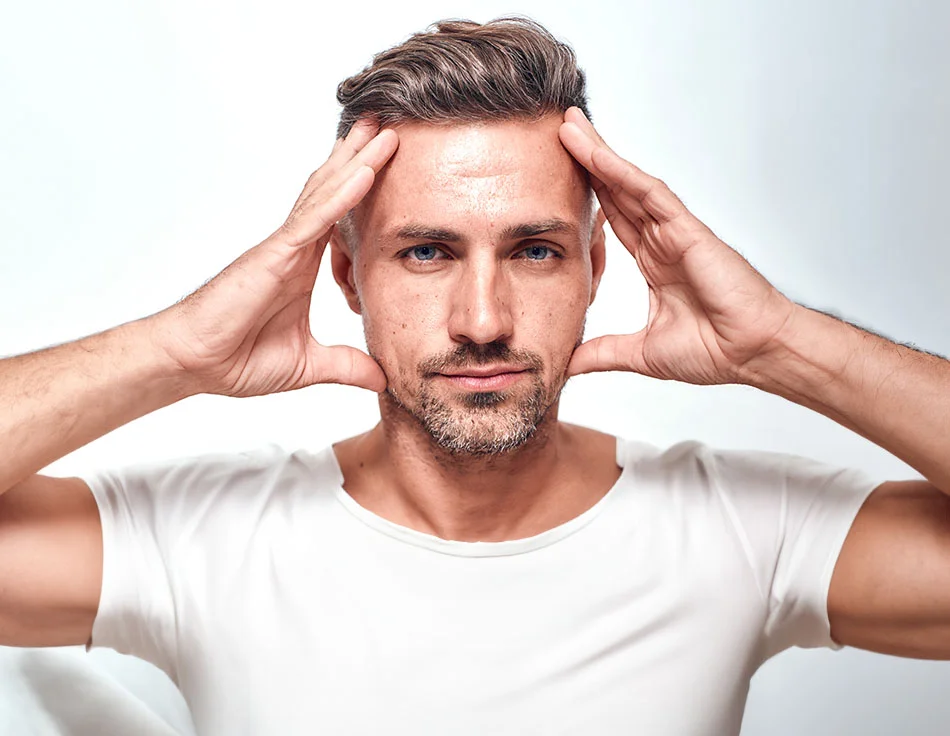 A man with light brown hair and a beard, wearing a white t-shirt, holding his hands on the sides of his face, looking directly at the camera against a white background - Brow Lift for men at HIM Plastic Surgery