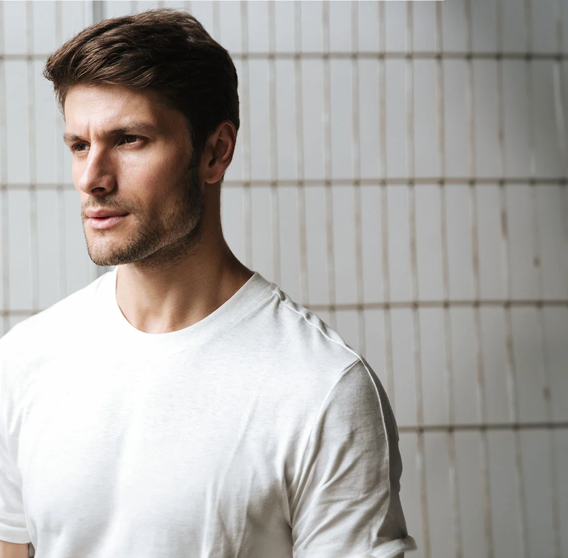 A man with short brown hair and a light beard, wearing a white t-shirt, standing indoors against a white tiled wall, looking off to the side - Buccal Fat Removal at HIM Plastic Surgery