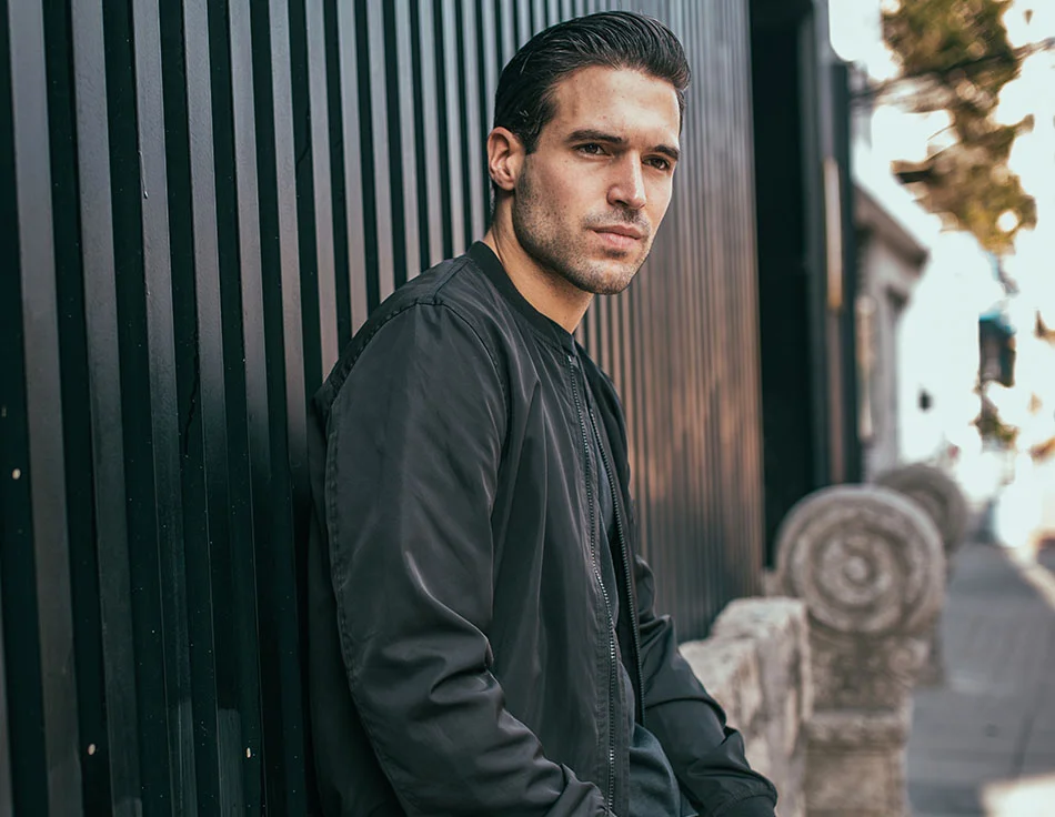 A man with dark slicked-back hair, wearing a black jacket, leaning against a black metal wall outdoors - Buccal Fat Removal at HIM Plastic Surgery