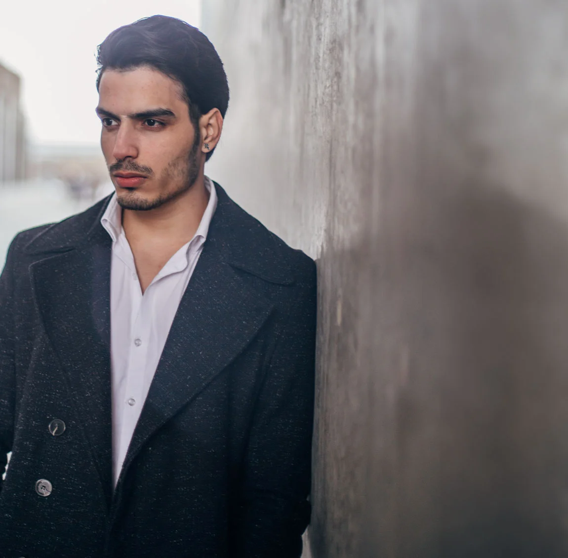 A man with dark hair and a beard, wearing a black coat and white shirt, leaning against a wall with a serious expression - Cheek Filler at HIM Plastic Surgery