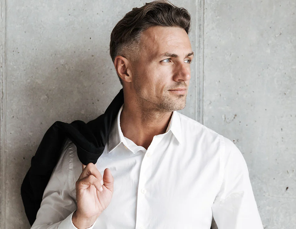 A man in a white shirt holding a black jacket over his shoulder, looking off to the side with a confident and relaxed demeanor - Cheek Filler at HIM Plastic Surgery