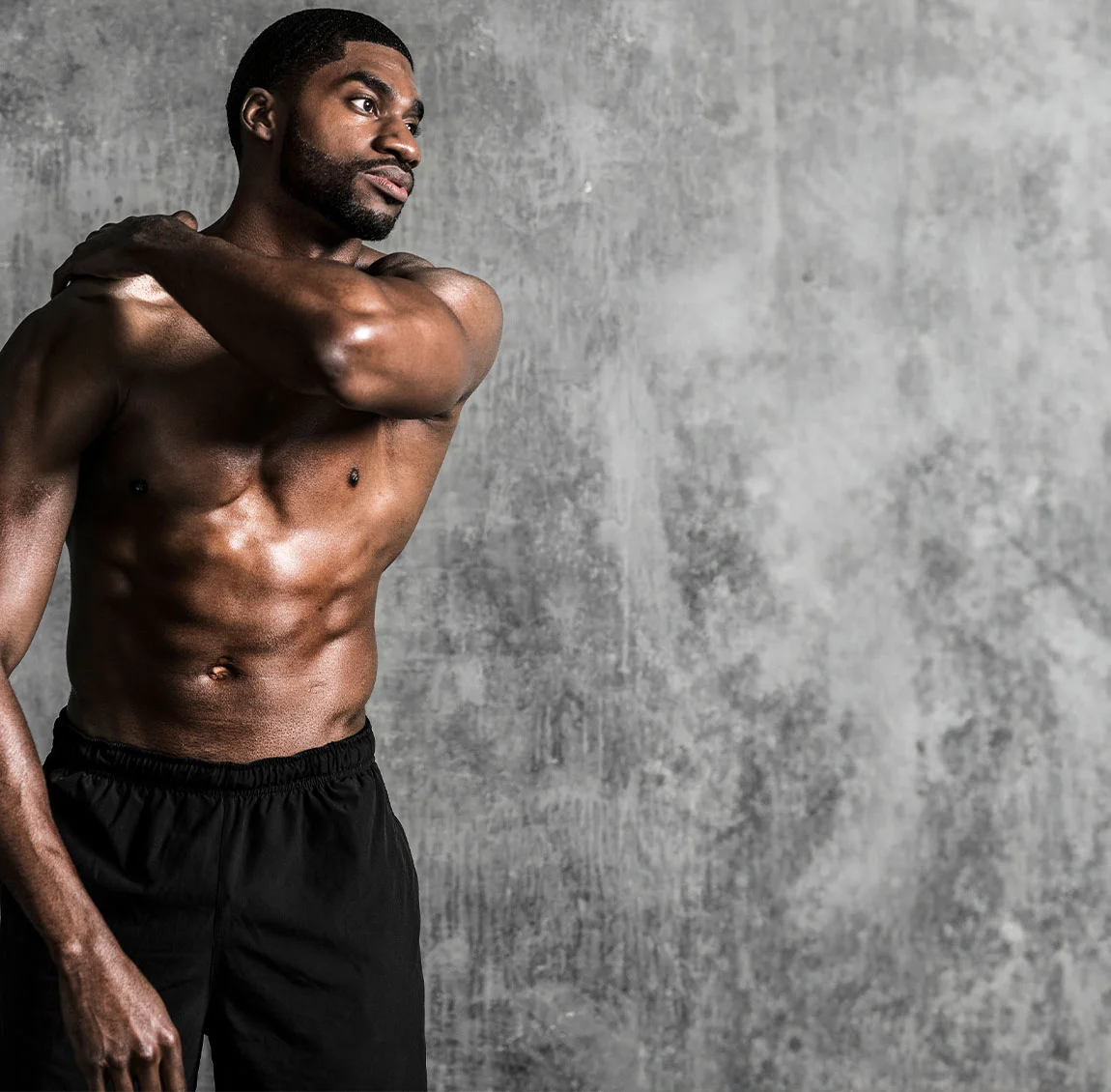 A shirtless man with a fit physique looking off into the distance, standing against a textured grey background, with one arm resting on his shoulder - Chest Wall Definition at HIM Plastic Surgery