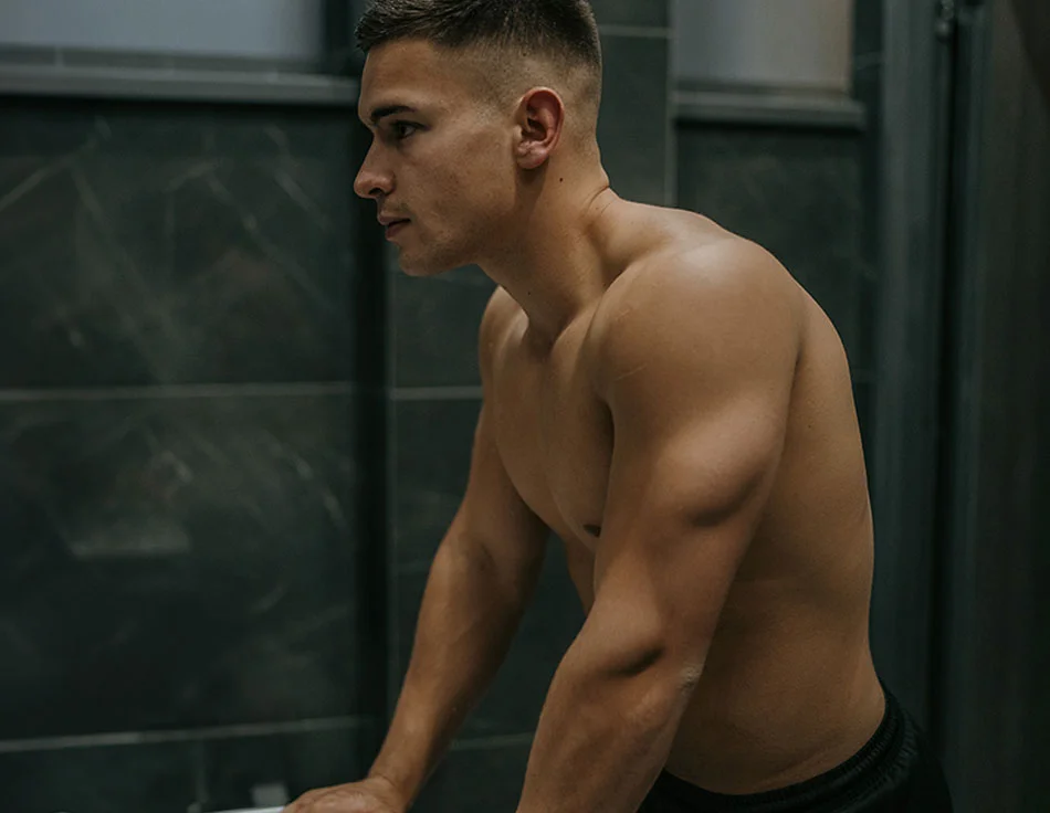 A shirtless man with a strong build leaning on a counter, looking off into the distance with a serious expression, set in a modern interior - Chest Wall Definition at HIM Plastic Surgery