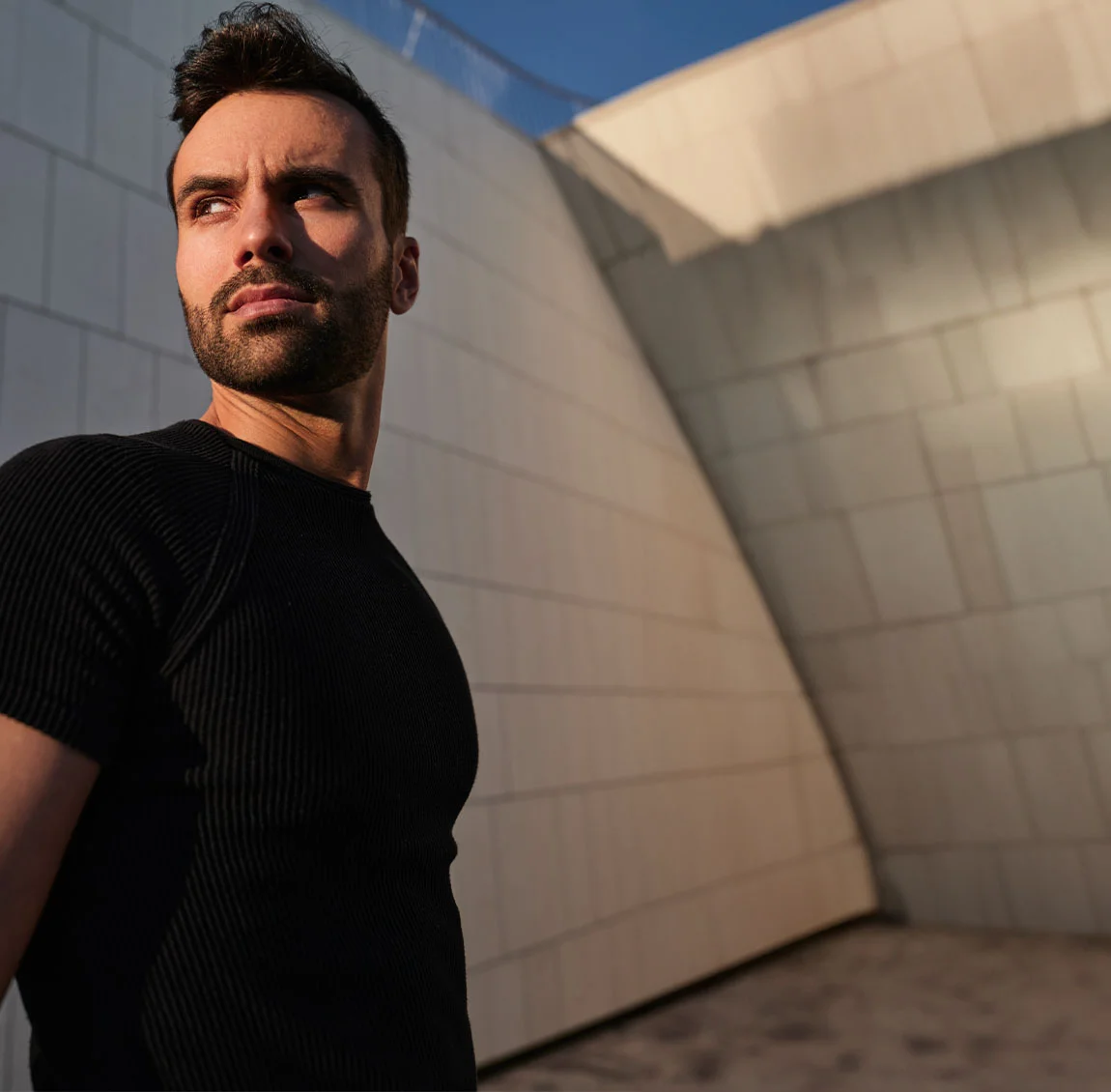 A man in a black t-shirt standing in the shadows of a modern building, looking off into the distance with a confident and contemplative expression - Chin Implant at HIM Plastic Surgery