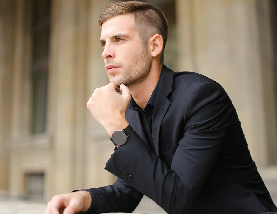 A man in a black suit resting his chin on his hand, looking thoughtful and reflective, set against a background of classical architecture - Chin Implant at HIM Plastic Surgery