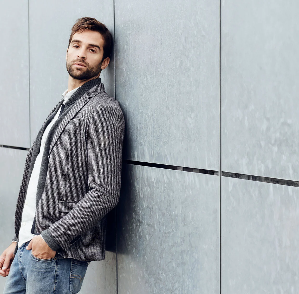 A man in a black coat and white shirt, leaning against a wall with a serious expression, appearing deep in thought - Daddy Do Over at HIM Plastic Surgery