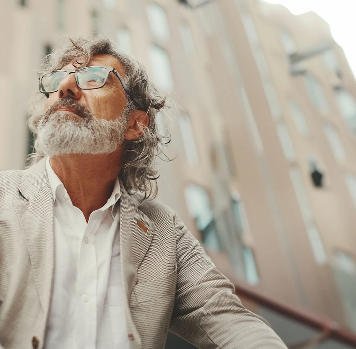 An older man with gray hair and a beard wearing glasses and a light suit, looking up - Eyelid Surgery at HIM Plastic Surgery