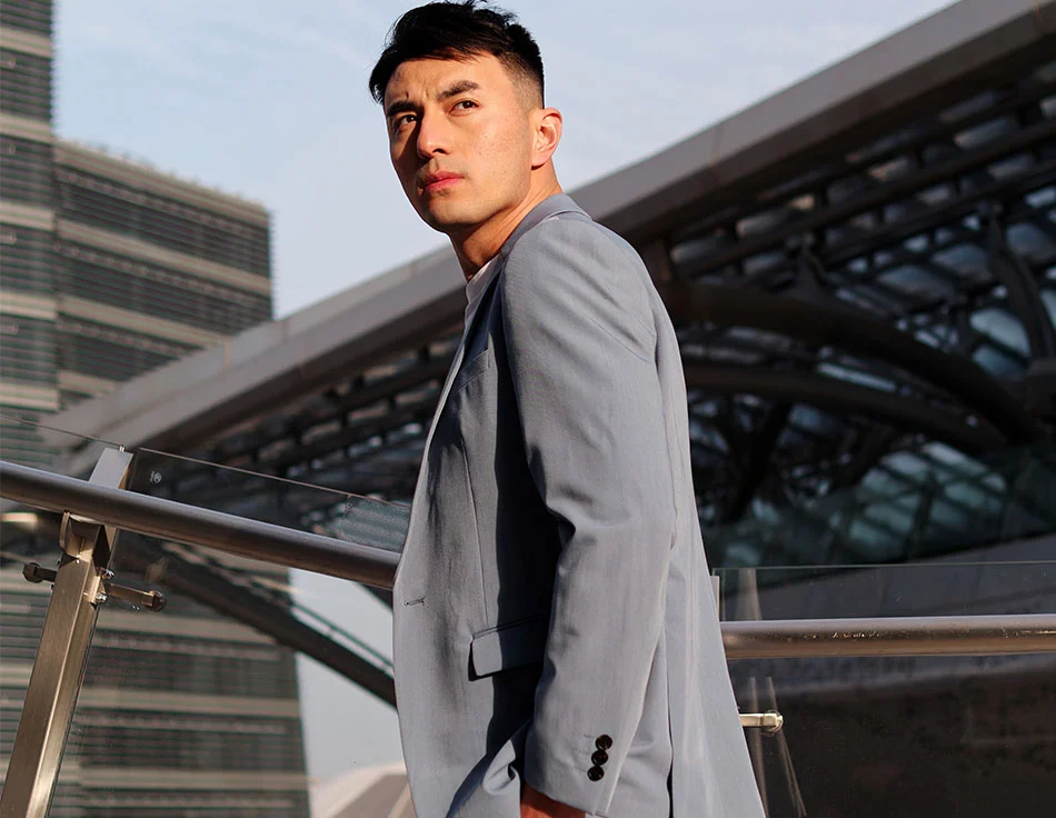 A man in a light blue suit standing on a balcony with a modern building in the background - Eyelid Surgery at HIM Plastic Surgery