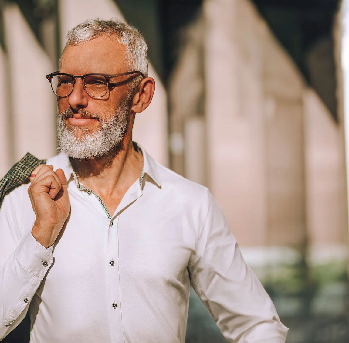 An older man with white hair and a beard wearing glasses and a white shirt, holding a jacket over his shoulder - Facelift at HIM Plastic Surgery