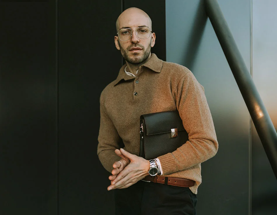 Bald man with glasses wearing a brown sweater and holding a black leather portfolio, posing confidently against a modern building - Functional Rhinoplasty at HIM Plastic Surgery