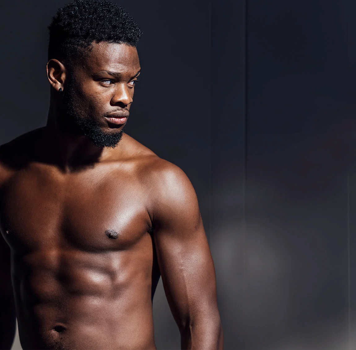 Athletic man with dark curly hair standing shirtless in dramatic lighting, highlighting his muscular physique - Gynecomastia after Steroid Use at HIM Plastic Surgery