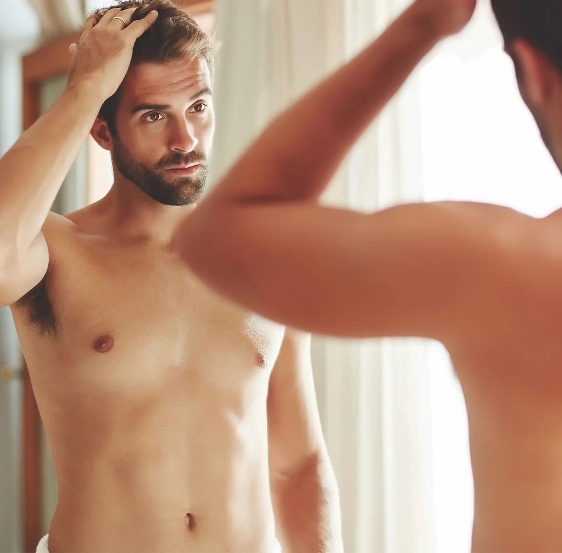 A man with a beard, looking at himself in the mirror, adjusting his hair, and wearing a white towel around his waist - Gynecomastia at HIM Plastic Surgery