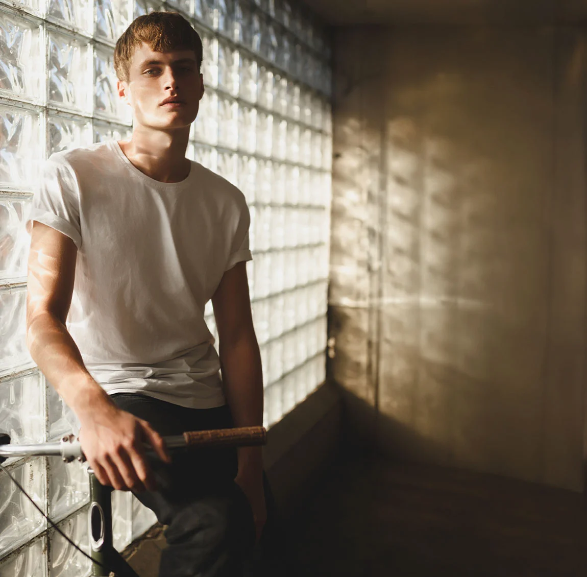 A young man with light brown hair, wearing a white t-shirt, sitting on a bicycle next to a wall made of glass bricks, with sunlight streaming in - Gynecomastia in Teens at HIM Plastic Surgery