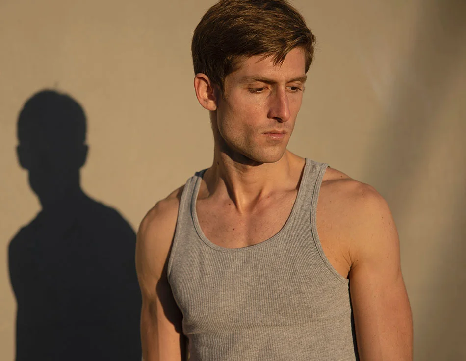 A man with short, light brown hair, wearing a grey tank top, standing against a beige wall with his shadow cast beside him - Gynecomastia at HIM Plastic Surgery