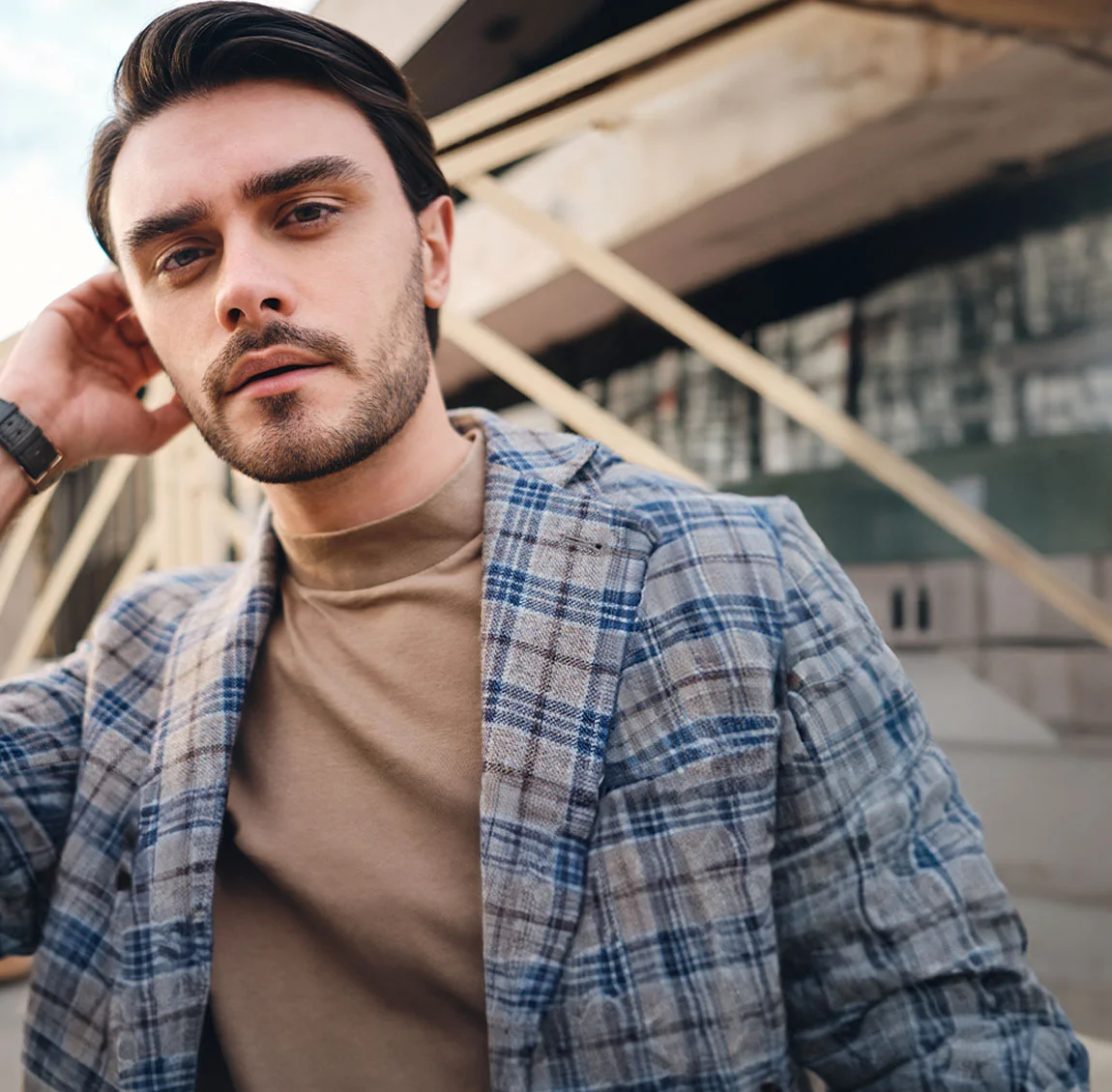 A man with dark hair, wearing a plaid jacket and beige t-shirt, standing outdoors in an urban setting, with his hand resting on his neck - Hairline Lowering at HIM Plastic Surgery