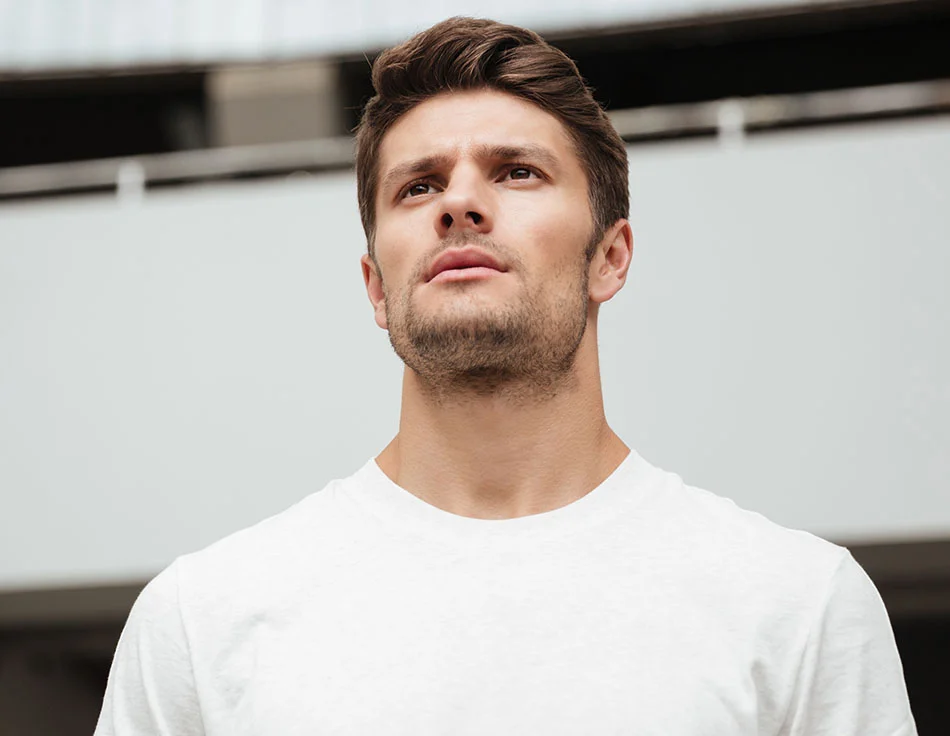 A confident man in a white t-shirt looking upwards with determination - Hairline Lowering at HIM Plastic Surgery