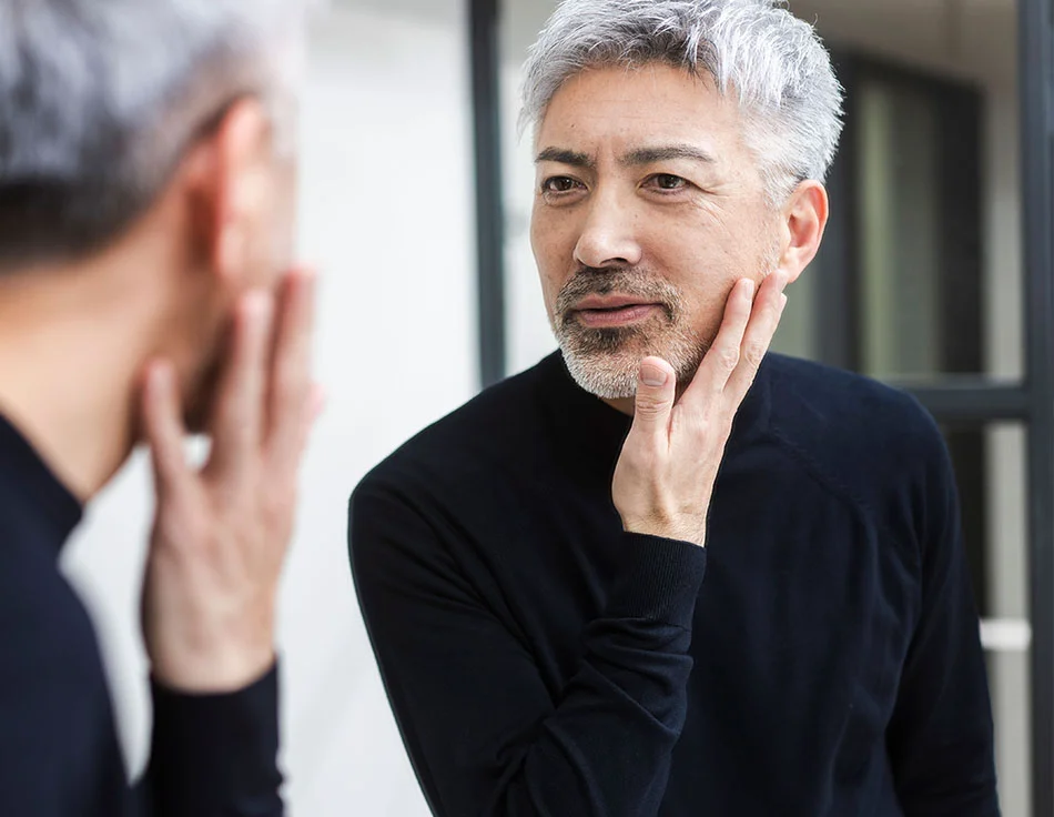 An older man with grey hair and beard looking at his reflection in the mirror and touching his face - Laser Skin Resurfacing at HIM Plastic Surgery