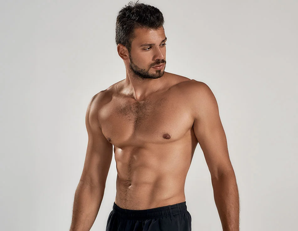 A shirtless, muscular man in black shorts looking to the side with a neutral expression - Liposuction at HIM Plastic Surgery