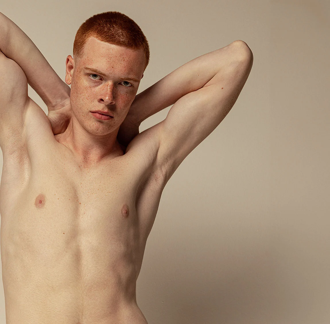 A red-haired, freckled young man with a determined look, standing shirtless with his arms raised behind his head, in front of a neutral background - Male Polands Syndrome Procedure at HIM Plastic Surgery