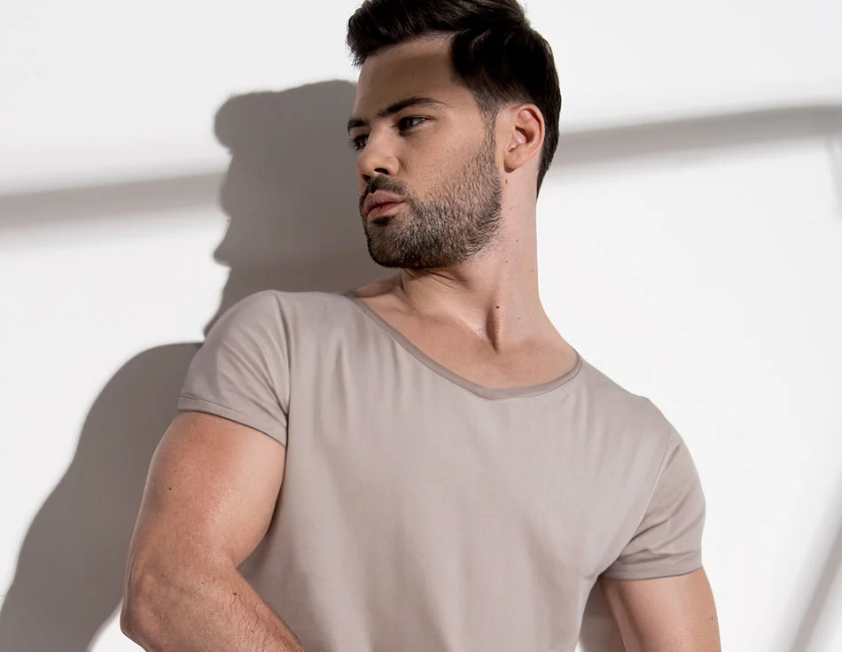 A man with a short beard and styled hair, wearing a beige t-shirt, looking to the side against a white wall with shadows - Male Polands Syndrome Procedure at HIM Plastic Surgery