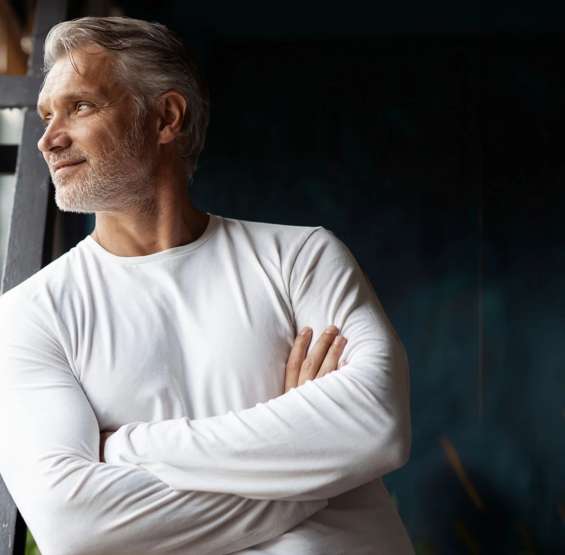 An older man with grey hair and a beard, wearing a white long-sleeve shirt, standing with his arms crossed, looking out thoughtfully in a dimly lit room - Neck and Chin Liposuction at HIM Plastic Surgery