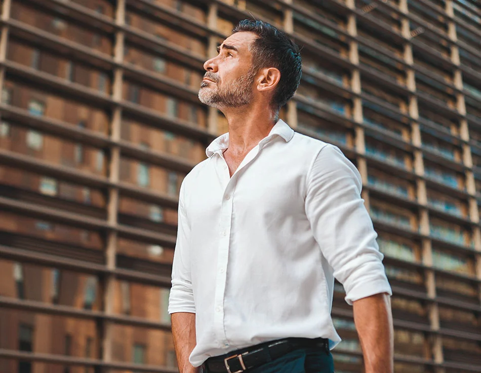 A mature man with a trimmed beard, wearing a white shirt, standing outside a building, looking up pensively - Neck and Chin Liposuction at HIM Plastic Surgery