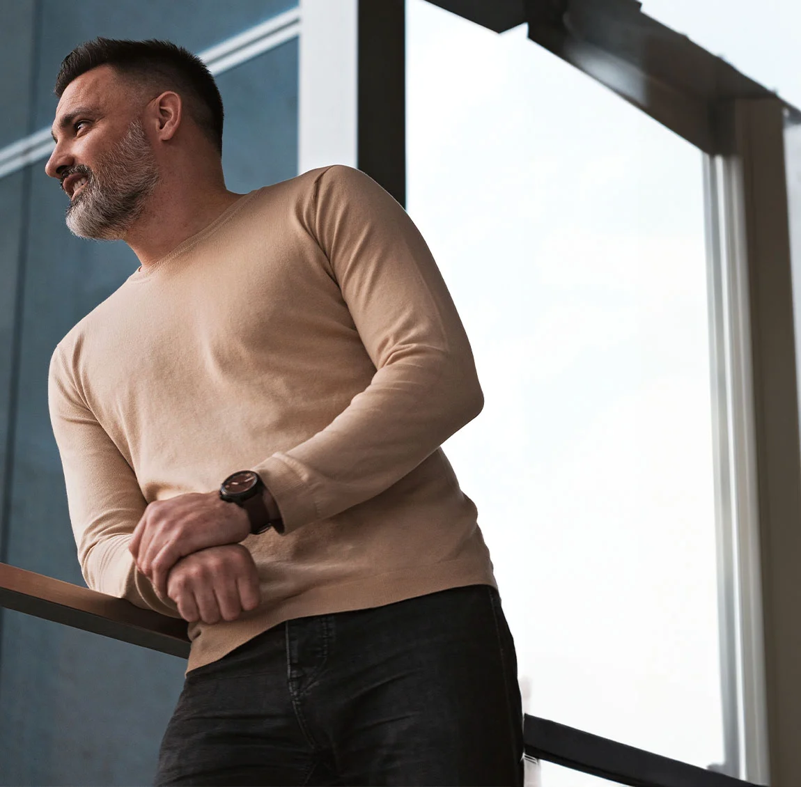 A man with neatly styled hair and a sharp jawline, wearing a white button-up shirt, standing against a concrete wall and looking off into the distance - Neck Lift at HIM Plastic Surgery