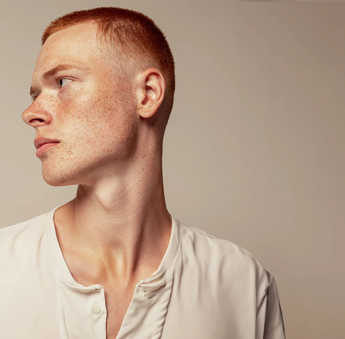 A man with short red hair and freckles, looking to the side against a tan background - Skin Care for Men at HIM Plastic Surgery