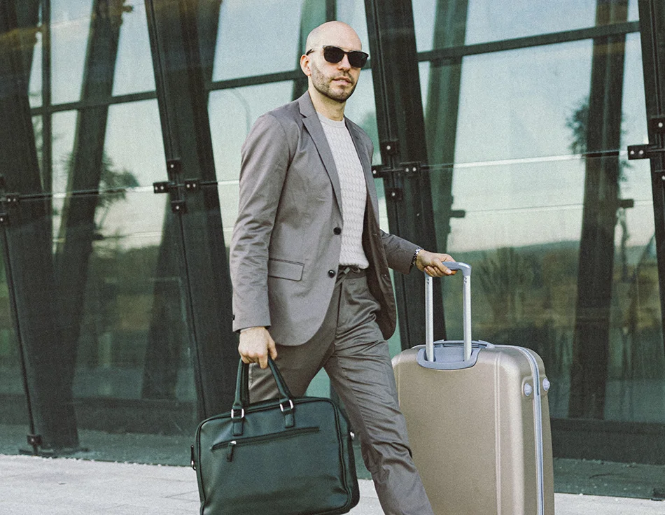 A man in a suit and sunglasses, walking with luggage - Out of Town Patient Resources at HIM Plastic Surgery