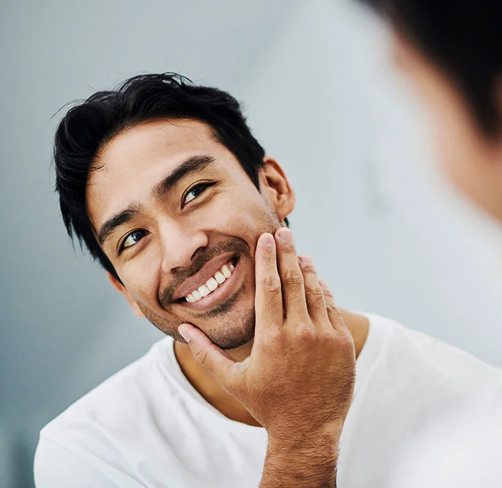 Man feeling his face, smiling while looking in the mirror | HIM Plastic Surgery Facial procedures